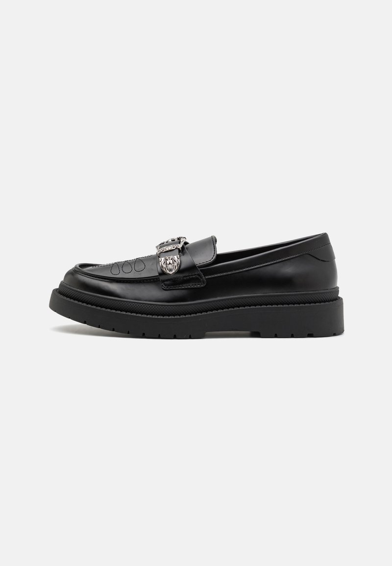 Mocassin en cuir noir avec semelle épaisse, boucle argentée décorative et détails de couture subtils sur un fond clair uni.