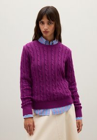 Lila Kabelstrickpullover mit geripptem Saum und Bündchen, über einem hellblauen gestreiften Hemd getragen, kombiniert mit einem neutralfarbenen Rock.