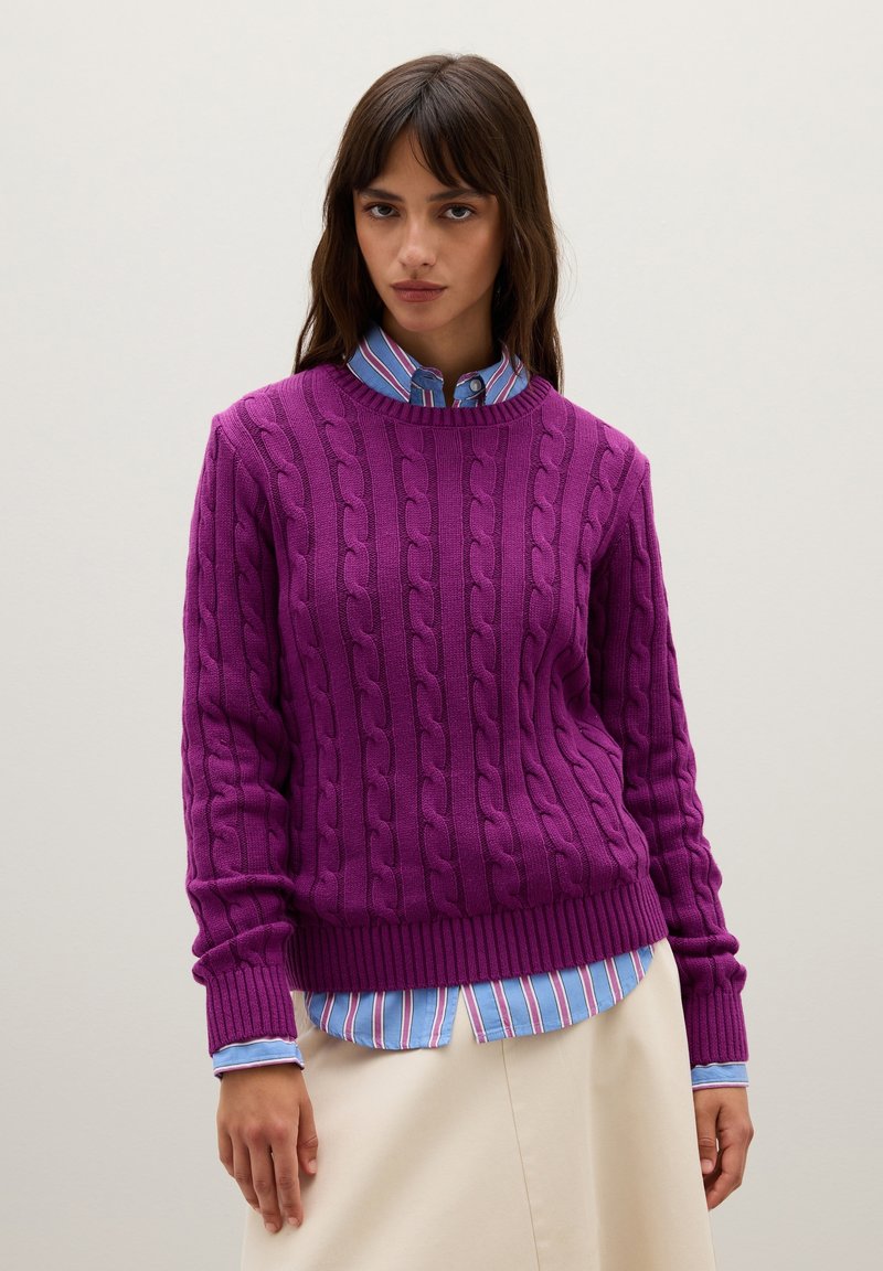 Lila Kabelstrickpullover mit geripptem Saum und Bündchen, über einem hellblauen gestreiften Hemd getragen, kombiniert mit einem neutralfarbenen Rock.
