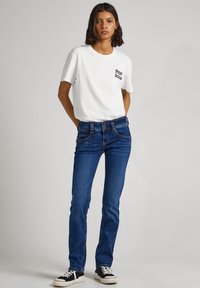 Mujer de pie con las manos en los bolsillos, vistiendo una camiseta blanca de Pepe Jeans, jeans azules y zapatillas a rayas en blanco y negro, frente a un fondo liso.