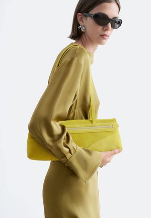 Vrouw met een zonnebril en een olijfgroene jurk draagt een bijpassende geelgroene handtas met ritssluiting over haar schouder.