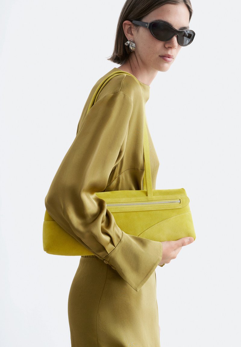 Mujer con gafas de sol y vestido verde oliva sostiene un bolso amarillo-verde a juego con detalle de cremallera sobre el hombro.