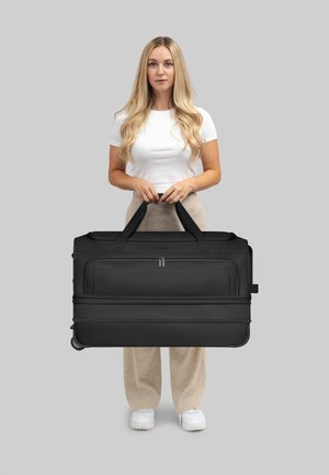 Sac de sport noir à roulettes avec double poignées, poche avant et texture de tissu lisse, se tenant debout sur un fond gris.