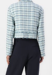 Vrouw in een kort, blauw-wit geblokt geweven jasje met rafelige zoom en marineblauwe high-waist broek, gezien vanuit de rug.