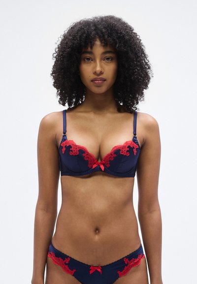 Soutien-gorge en dentelle bleu marine avec accents floraux rouges, armatures, bretelles réglables et petit nœud au centre. Bas bikini assortis.