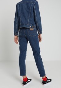 Mörk denimjacka och jeans med slim fit, med fickor och en brun lapp på ryggen. Svarta sneakers med vita detaljer och röda strumpor.