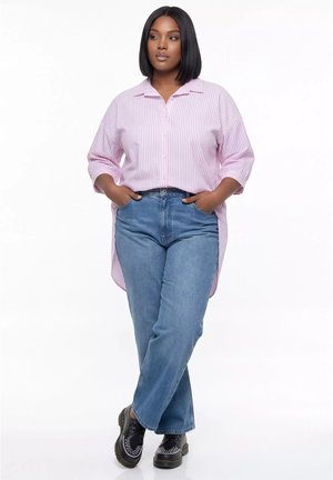 Kobieta stoi z rękami w kieszeniach, ubrana w różową, paskowaną długą koszulę, niebieskie dżinsy i czarne buty na białym tle.