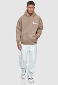Brun hettegenser med frontlomme og hvit "Dropsize"-tekst, kombinert med lyseblå vide jeans og hvite sneakers med svarte detaljer.