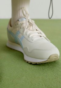 Sneaker bianco con accenti blu chiaro, parte superiore testurizzata e suola beige. Presenta lacci, collare imbottito e un design atletico curvato.
