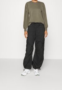 Camiseta de manga larga verde oliva con hombros abullonados, combinada con pantalones cargo negros de corte holgado. Zapatillas deportivas blancas con detalles en gris.
