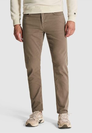 Άνδρας που φοράει καφέ παντελόνια slim-fit συνδυασμένα με μπεζ και λευκά αθλητικά παπούτσια, στέκεται μπροστά σε απλό ανοιχτό φόντο.