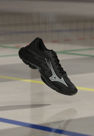Chaussure de sport noire avec un logo blanc et un texte en suspension au-dessus d'un terrain de sport avec des lignes multicolores.