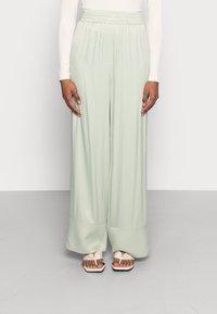 Femme portant un pantalon large vert menthe avec une taille élastique et des sandales ouvertes blanches, se tenant devant un fond uni.