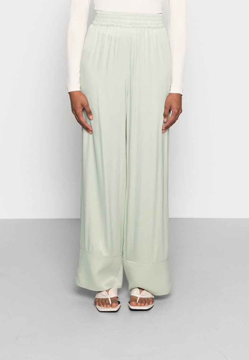 Femme portant un pantalon large vert menthe avec une taille élastique et des sandales ouvertes blanches, se tenant devant un fond uni.