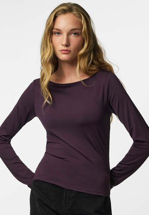 Jeune femme aux longs cheveux blonds portant un haut à manches longues violet foncé et un pantalon noir, debout, les mains sur les hanches.