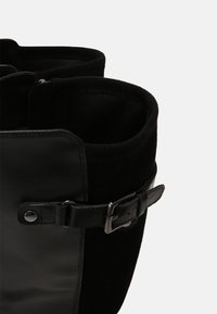 Botte en daim noir et cuir avec fermeture à boucle. Présente un sommet arrondi, une texture lisse et des matériaux contrastés pour un intérêt visuel.