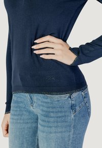 Maglia a maniche lunghe blu navy con texture a coste, dettagli argentati ai polsini e all'orlo, con logo in basso, abbinata a jeans blu chiaro.