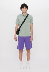 Svetlozelená polo tričko s logom EA7, skombinované s fialovými šortkami. Model má na sebe biele tenisky a čiernu crossbody tašku. Jednoduché pozadie.