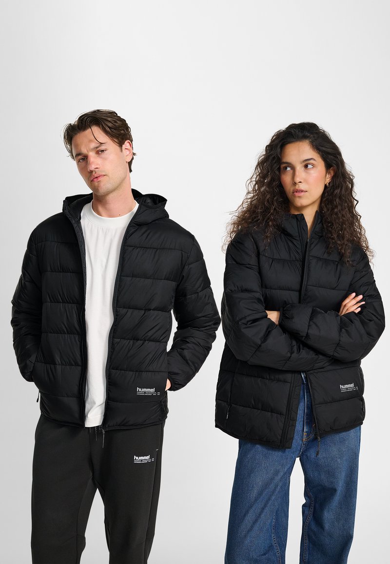 Man en vrouw dragen zwarte donsjassen; man met handen in zakken, vrouw met armen gekruist; man draagt zwarte broek, vrouw draagt blauwe spijkerbroek.