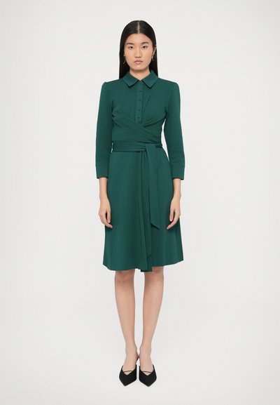 Groene wikkeljurk met knoopsluiting, lange mouwen en ceintuurdetaillering. Soepele stof, tot op de knieën, gecombineerd met zwarte puntige schoenen.