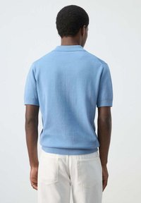 Hombre de pie de espaldas, vistiendo una camiseta tipo polo de punto azul claro de manga corta y pantalones blancos contra un fondo liso.