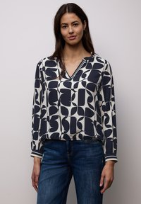 Femme portant un chemisier à manches longues à motif abstrait noir et blanc et un jean bleu, debout devant un fond uni clair.