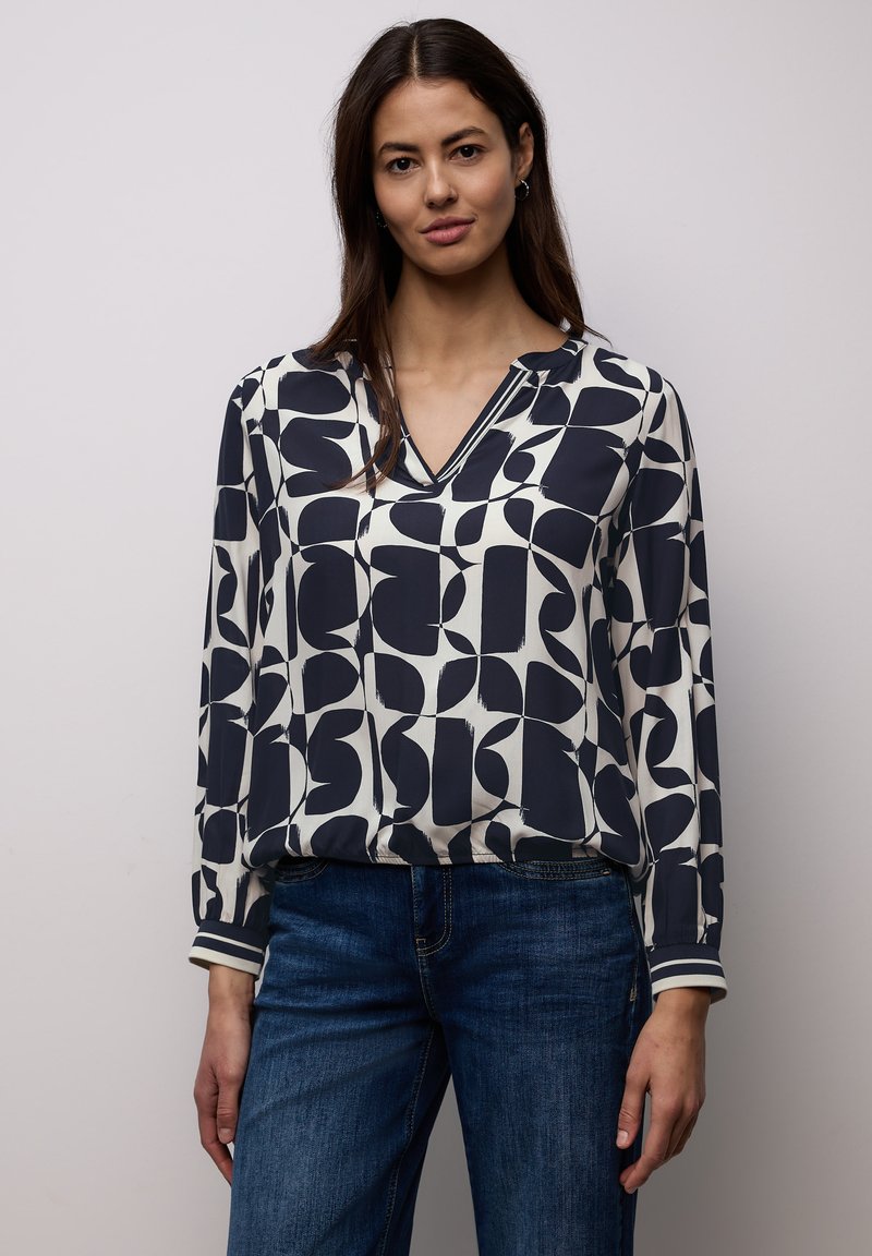 Femme portant un chemisier à manches longues à motif abstrait noir et blanc et un jean bleu, debout devant un fond uni clair.