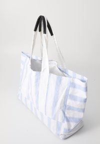 Grande bolsa tote feita de tecido branco com listras azuis. Apresenta duas alças de lona brancas e um detalhe em couro preto na parte superior.