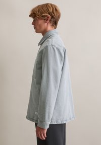 Hellblaue Jeansjacke mit langen Ärmeln, Kragen, Brusttasche und geradem Schnitt. Weiche Textur, minimale Nahtdetails.