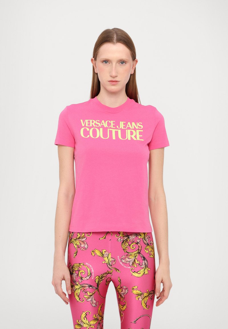 Roza bombažna majica s logotipom "VERSACE JEANS COUTURE" v rumenem tisku. Model nosi vzorčaste roza legice z rumenimi akcenti.