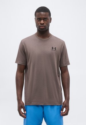 Młody mężczyzna z krótkimi włosami, ubrany w brązowy T-shirt Under Armour i jaskrawo niebieskie szorty, stoi na tle jednolitego białego tła.