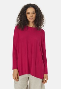 T-shirt ample à manches longues en rose vif, fabriqué en tissu doux avec des détails côtelés aux manches, porté avec un pantalon léger et texturé.