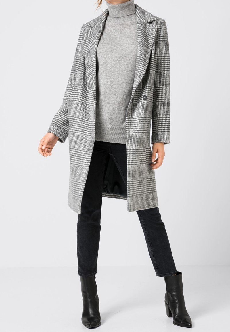 Grå rutiga kappa med slagkrage, dubbelknäppta knappar, långa ärmar och en avslappnad passform över en grå turtleneck och svarta jeans.