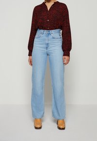 Kvinna i högmidjade ljusblå raka jeans, brun blommig skjorta med knappar och beige klackboots mot vit bakgrund.