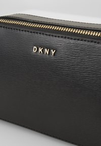 Černá texturovaná kosmetická taštička z umělé kůže s viditelným zlatým zipem. Na hladkém černém povrchu se nachází zlaté logo DKNY. Kompaktní a obdélníkový design.