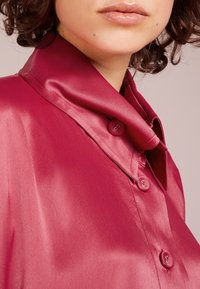 Lesklá, saténová bordeaux blůza s drapovaným límcem a zapínáním na dva knoflíky, která ukazuje hladkou texturu a jemný lesk.