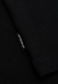 Gros plan sur un tissu noir avec une petite étiquette noire cousue affichant le logo blanc "ombre" et quatre barres verticales.