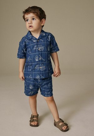 Jauns zēns skatās uz augšu, valkā zilu īsroku krekls un atbilstošas šortus ar baltu jūras dzīves rakstu, stāvot uz bēša grīdas.