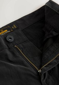 Gros plan sur un pantalon noir dézippé avec un bouton-pression et une étiquette de marque visible à l'intérieur de la ceinture.