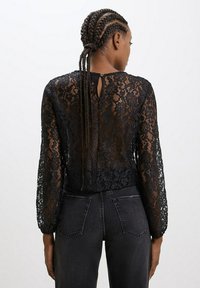 Blusa nera in pizzo con maniche lunghe, caratterizzata da uno scollo a chiave sul retro e un motivo floreale texture. Abbinata a pantaloni in denim scuro.