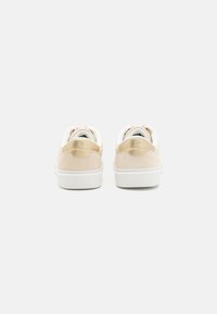 Une paire de baskets beige et blanche avec des accents dorés vues de dos sur un fond blanc.
