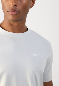 Lichtgrijs katoenen t-shirt met een ronde halslijn. Heeft een klein wit logo; zachte textuur en korte mouwen. Minimalistisch ontwerp.