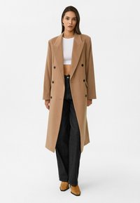 Lang, dubbel-breasted tan coat met notch lapels, twee voorpockets en donkere knopen, gedragen over een witte crop top en zwarte wijde broek.