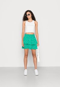 Top cropped branco, saia verde em camadas com padrão floral e sapatilhas brancas. Modelo segurando uma pequena bolsa branca, usando óculos de sol.