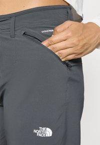 Ręka wsuwająca się do zapinanej na zamek kieszeni szarych spodni The North Face WindWall, noszonych z widocznym białym rękawem.