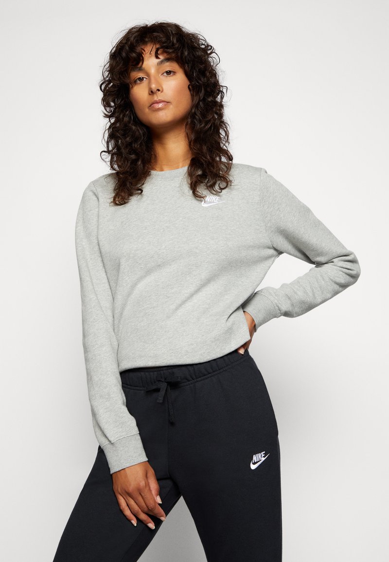 Sweatshirt gris à manches longues et col rond, arborant un petit logo Nike blanc sur la poitrine. Associé à un pantalon de survêtement noir.