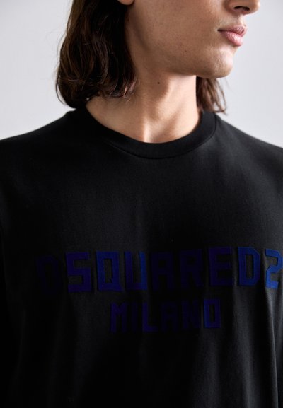 Noor inimene, kellel on õlavarrepikkune juuksed, kannab musta särki, millel on sinine tekst "DSQUARED2 MILANO", seistes paremale puutumatul taustal.