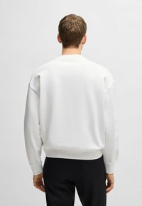 Fehér pulóver laza szabással, bordás mandzsettákkal és szegéllyel. A hátán egy kis folt található, amely további részletet ad. Sima textúra, minimális design.