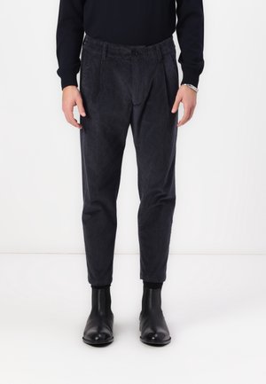 Hombre con pantalones de pana gris oscuro, un suéter negro y botines de cuero negro, de pie frente a un fondo blanco.