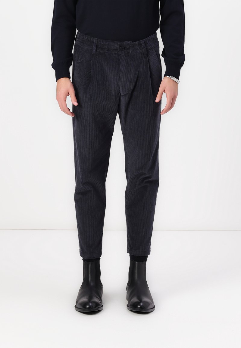 Homme portant un pantalon en velours côtelé gris foncé avec un pull noir et des bottines en cuir noir, debout sur un fond blanc.